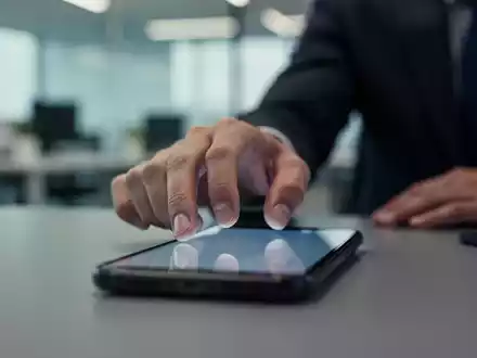 Eine Hand greift nach einem Smartphone in einem unscharfen Büroumfeld, das die ständige Erreichbarkeit symbolisiert. - Foto: über boerse-global.de