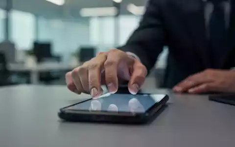 Eine Hand greift nach einem Smartphone in einem unscharfen Büroumfeld, das die ständige Erreichbarkeit symbolisiert. - Foto: über boerse-global.de