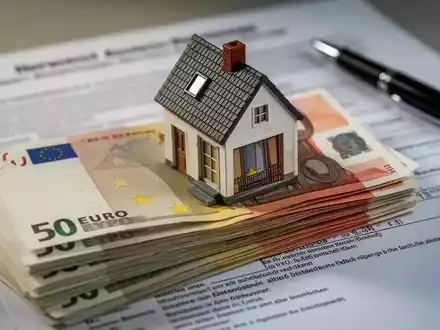 Ein Stapel deutscher Euro-Banknoten, teilweise verdeckt von einem Miniatur-Hausmodell, mit einem verschwommenen Rechtsdokument im Hintergrund. - Foto: über boerse-global.de