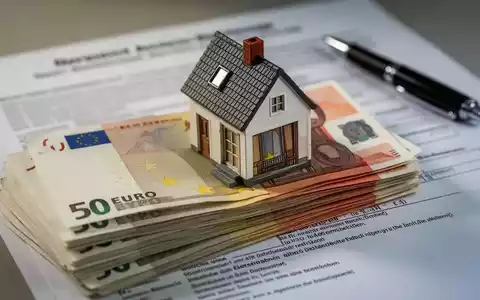 Ein Stapel deutscher Euro-Banknoten, teilweise verdeckt von einem Miniatur-Hausmodell, mit einem verschwommenen Rechtsdokument im Hintergrund. - Foto: über boerse-global.de
