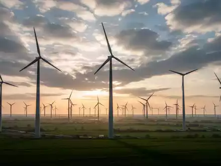 Ein weitläufiger Windpark mit hohen Windturbinen im Sonnenuntergang, vor einem dramatischen Himmel. - Foto: über boerse-global.de