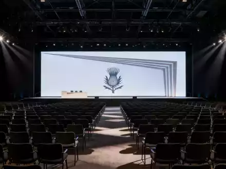Eine moderne, leere Konferenzhalle mit Reihen von Stühlen, die auf eine beleuchtete Bühne blicken, bereit für eine Hauptversammlung. - Foto: über boerse-global.de
