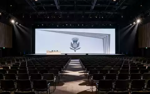 Eine moderne, leere Konferenzhalle mit Reihen von Stühlen, die auf eine beleuchtete Bühne blicken, bereit für eine Hauptversammlung. - Foto: über boerse-global.de