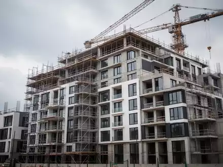 Ein unfertiges modernes Wohngebäude mit Baugerüsten und einem Kran vor einem bewölkten Himmel, das die Herausforderungen im Wohnungsbau darstellt. - Foto: über boerse-global.de