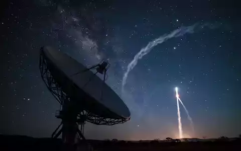 Eine Satellitenschüssel vor einem Sternenhimmel mit einer angedeuteten Raketenspur in der Ferne. - Foto: über boerse-global.de