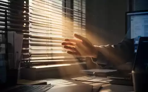 Eine Hand greift nach einem Sonnenstrahl, der durch Jalousien in ein dunkles Büro fällt, Symbol für Vitamin-D-Mangel. - Foto: über boerse-global.de