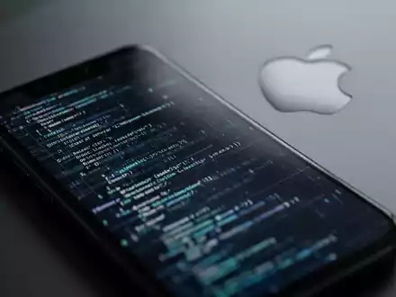 Close-up of an iPhone screen showing abstract code, with a subtle Apple logo in the background, symbolizing software updates. - Foto: über boerse-global.de