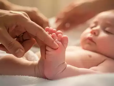 Eine Nahaufnahme eines Babyfußes, auf dem ein Finger sanft einen Akupressurpunkt stimuliert, mit unscharfen Erwachsenenhänden im Hintergrund. - Foto: über boerse-global.de