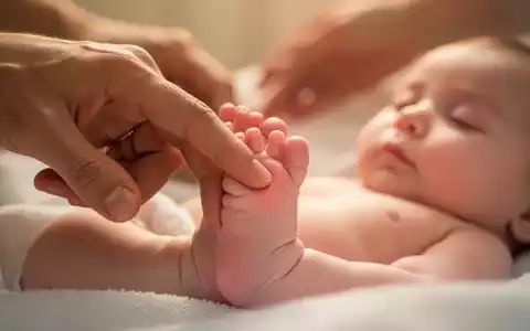 Eine Nahaufnahme eines Babyfußes, auf dem ein Finger sanft einen Akupressurpunkt stimuliert, mit unscharfen Erwachsenenhänden im Hintergrund. - Foto: über boerse-global.de