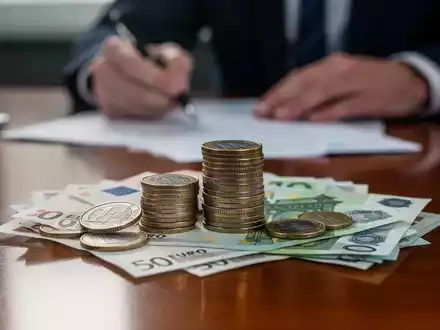 Ein Stapel deutscher Euro-Münzen und -Banknoten auf einem Schreibtisch, unscharfer Hintergrund zeigt eine Person, die ein Dokument unterschreibt. - Foto: über boerse-global.de