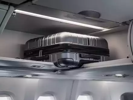 Eine Nahaufnahme eines eleganten Handgepäckstücks, das in ein Flugzeug-Gepäckfach geschoben wird, mit weichem, diffusem Licht. - Foto: über boerse-global.de