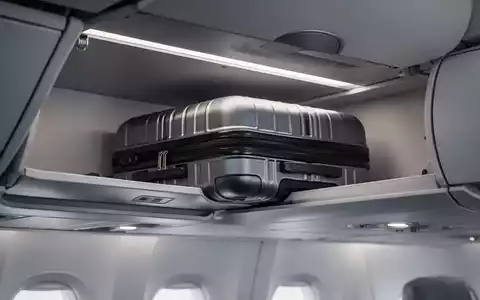 Eine Nahaufnahme eines eleganten Handgepäckstücks, das in ein Flugzeug-Gepäckfach geschoben wird, mit weichem, diffusem Licht. - Foto: über boerse-global.de