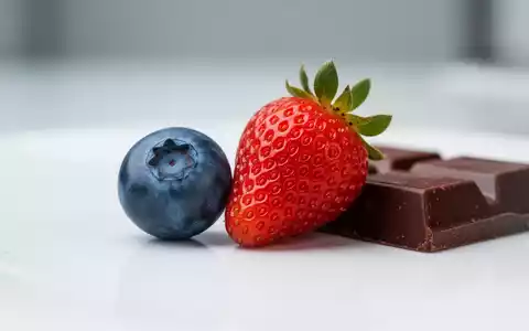 Nahaufnahme von frischen Blaubeeren, einer Erdbeere und einem Stück dunkler Schokolade auf einer weißen Oberfläche. - Foto: über boerse-global.de