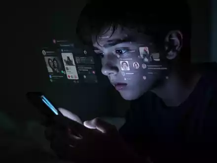 Eine junge Person, im Profil, blickt auf ein leuchtendes Smartphone in einem dunklen Raum, was Angst und Isolation symbolisiert. - Foto: über boerse-global.de