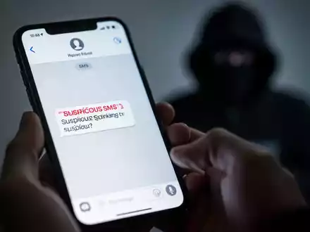 Nahaufnahme eines Smartphones mit einer verdächtigen SMS. Im Hintergrund verschwommen die Silhouette eines Hackers, der digitale Bankbetrügereien symbolisiert. - Foto: über boerse-global.de