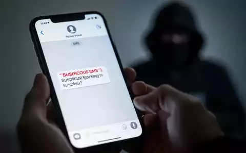 Nahaufnahme eines Smartphones mit einer verdächtigen SMS. Im Hintergrund verschwommen die Silhouette eines Hackers, der digitale Bankbetrügereien symbolisiert. - Foto: über boerse-global.de