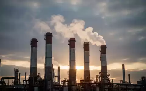 Industrieschornsteine emittieren Dampf oder weißen Rauch, vor einem dramatischen Himmel, der regulatorische Änderungen und wirtschaftliche Auswirkungen symbolisiert. - Foto: über boerse-global.de
