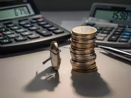 Ein Stapel Euro-Münzen mit einem Taschenrechner und einem Stift im Hintergrund, der steigende Kosten symbolisiert. - Foto: über boerse-global.de