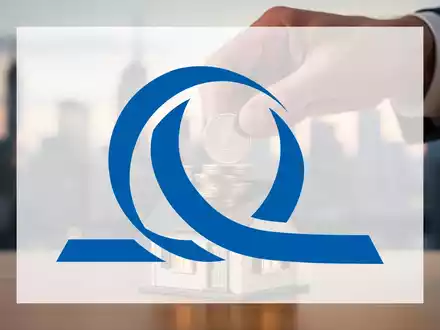 Eine Hand platziert Euro-Münzen auf einem Miniaturhaus, mit verschwommener Finanzstadt im Hintergrund. - Foto: über boerse-global.de