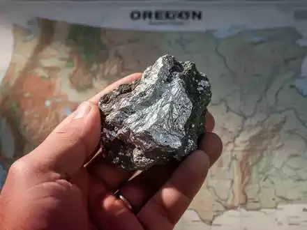 Nahaufnahme einer Hand, die ein rohes Silbererzmuster hält, mit einer verschwommenen geologischen Karte von Oregon im Hintergrund. - Foto: über boerse-global.de