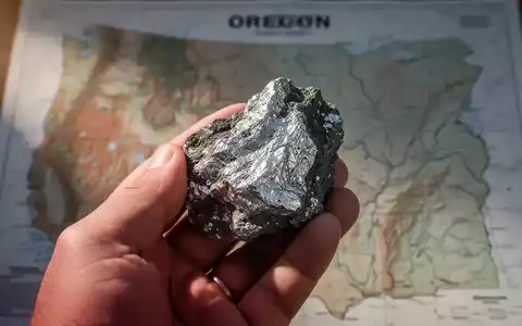 Nahaufnahme einer Hand, die ein rohes Silbererzmuster hält, mit einer verschwommenen geologischen Karte von Oregon im Hintergrund. - Foto: über boerse-global.de