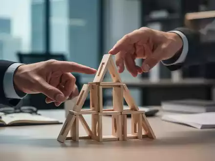 Eine Hand berührt vorsichtig ein Kartenhaus, das persönliche Ziele und Errungenschaften symbolisiert, in einem modernen Büro. - Foto: über boerse-global.de