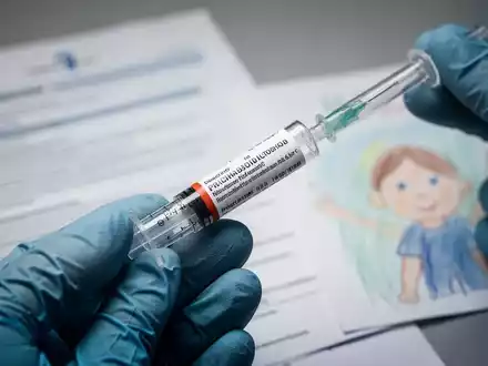 Eine Nahaufnahme einer vorgefüllten Spritze, die von einer behandschuhten Hand gehalten wird, mit medizinischen Dokumenten und einer Kinderzeichnung im Hintergrund. - Foto: über boerse-global.de