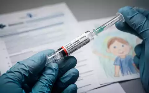 Eine Nahaufnahme einer vorgefüllten Spritze, die von einer behandschuhten Hand gehalten wird, mit medizinischen Dokumenten und einer Kinderzeichnung im Hintergrund. - Foto: über boerse-global.de