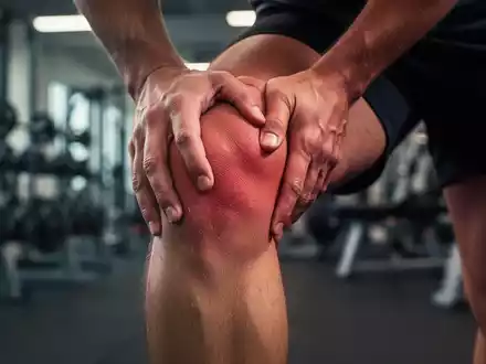 Nahaufnahme eines Knies, das leichte Schmerzen anzeigt, mit verschwommenem Hintergrund eines Fitnessstudios. - Foto: über boerse-global.de