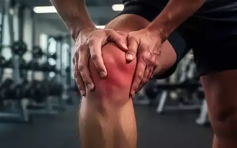 Nahaufnahme eines Knies, das leichte Schmerzen anzeigt, mit verschwommenem Hintergrund eines Fitnessstudios. - Foto: über boerse-global.de