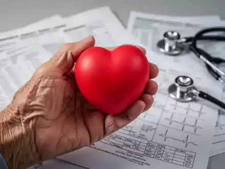 Ăltere Hand hĂ€lt einen herzförmigen Stressball; im Hintergrund verschwommen medizinische Diagramme und ein Stethoskop. - Foto: ĂŒber boerse-global.de