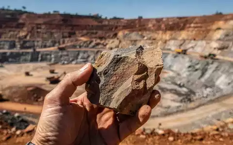 Geologenhand hält eine Gesteinsprobe mit einem Bergbaugelände im Hintergrund, das die Erkundung seltener Erden darstellt. - Foto: über boerse-global.de