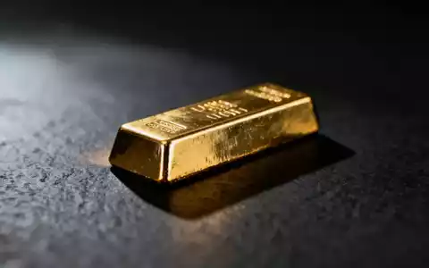 Ein einzelner glänzender Goldbarren ruht auf einer dunklen, texturierten Oberfläche, teilweise im Schatten. - Foto: über boerse-global.de