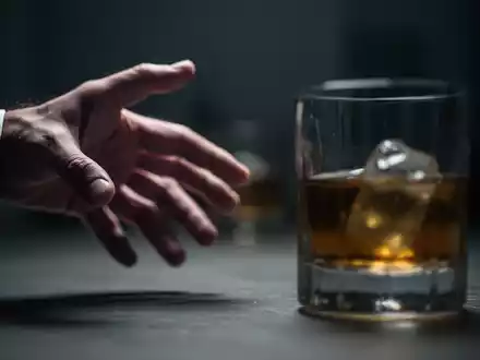 Eine einzelne Hand streckt sich unscharf nach einem Glas Alkohol aus, im Hintergrund verschwommen und dunkel. - Foto: über boerse-global.de