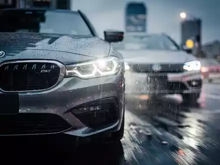 Ein BMW-Kühlergrill spiegelt die Scheinwerfer eines VW, auf einer nassen Straße bei Dämmerung, symbolisiert Wettbewerb. - Foto: über boerse-global.de