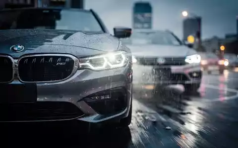 Ein BMW-Kühlergrill spiegelt die Scheinwerfer eines VW, auf einer nassen Straße bei Dämmerung, symbolisiert Wettbewerb. - Foto: über boerse-global.de
