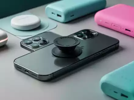 A modern smartphone with a low-profile PopSockets grip, surrounded by colorful wireless chargers and power banks on a clean surface. - Foto: über boerse-global.de