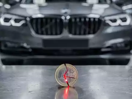 Ein gebrochenes Euro-Stück liegt vor dem Kühlergrill eines modernen BMW, im Hintergrund eine Fabrikhalle. Symbolisiert sinkenden Cashflow. - Foto: über boerse-global.de