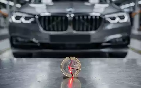 Ein gebrochenes Euro-Stück liegt vor dem Kühlergrill eines modernen BMW, im Hintergrund eine Fabrikhalle. Symbolisiert sinkenden Cashflow. - Foto: über boerse-global.de