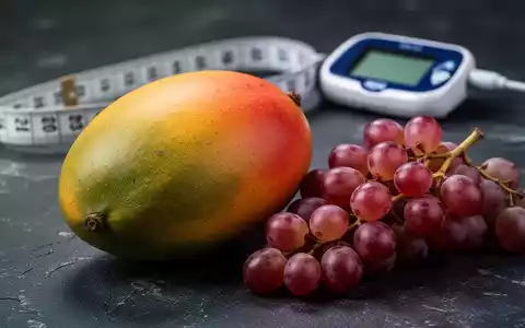 Nahaufnahme einer reifen Mango und Trauben auf dunklem Hintergrund, mit einem unscharfen Blutzuckermessgerät im Hintergrund. - Foto: über boerse-global.de
