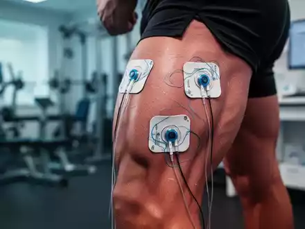 Nahaufnahme eines Oberschenkels mit TENS-Elektroden, die auf die Haut geklebt sind, im Hintergrund verschwommen ein Fitnessstudio. - Foto: über boerse-global.de