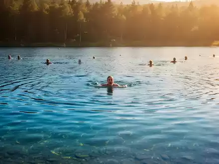 Ein ruhiger, klarer See bei Sonnenuntergang mit badenden Menschen in der Ferne, der die Sommerhitze und die Gefahren beim Schwimmen symbolisiert. - Foto: über boerse-global.de