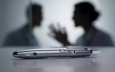 Eine Nahaufnahme eines modernen Injektionspens mit unscharfem Hintergrund, der eine Person andeutet, die den Stift betrachtet. - Foto: über boerse-global.de
