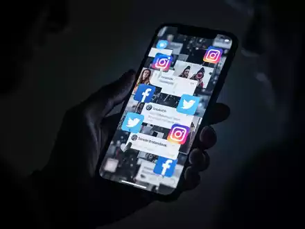 A shadowy hand holds a smartphone displaying distorted social media icons, symbolizing tech-enabled abuse and digital threats. - Foto: über boerse-global.de