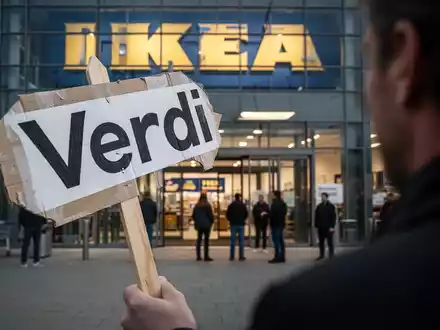 Ein Protestschild mit 'Verdi' davor, dahinter unscharf ein großes Möbelgeschäft mit versammelten Menschen, die einen Streik andeuten. - Foto: über boerse-global.de