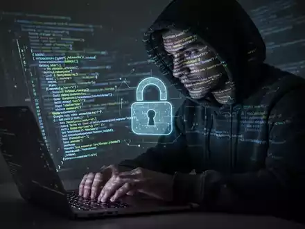 Eine Person im Kapuzenpulli tippt auf einem Laptop, mit leuchtenden Codezeilen, die ihr Gesicht und den Bildschirm beleuchten, symbolisiert Cyberbedrohungen. - Foto: über boerse-global.de