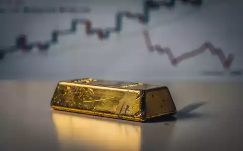 Ein leicht getrübter Goldbarren spiegelt Schatten wider, vor einem unscharfen Hintergrund mit fallenden Finanzdiagrammen. - Foto: über boerse-global.de