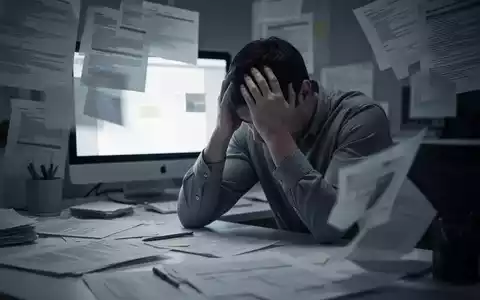 Eine Person sitzt am Schreibtisch, den Kopf in den Händen, umgeben von verschwommenen Dokumenten und einem leuchtenden Computerbildschirm. - Foto: über boerse-global.de
