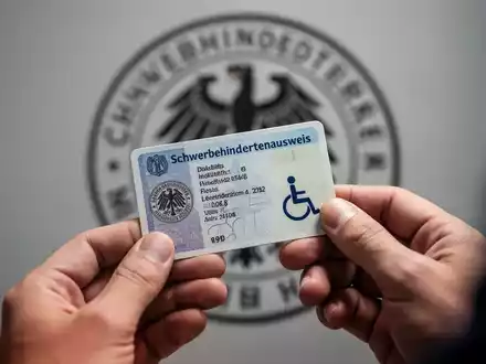 Hände halten einen deutschen Schwerbehindertenausweis, im Hintergrund verschwommenes Behördensiegel, das Verwaltungsprozesse andeutet. - Foto: über boerse-global.de