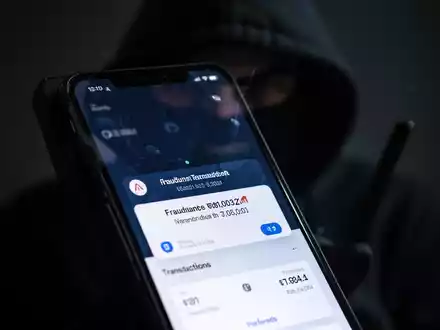 Ein Smartphone zeigt eine gefälschte Banking-App mit einer Betrugswarnung; im unscharfen Hintergrund ist schemenhaft ein Betrüger zu sehen. - Foto: über boerse-global.de